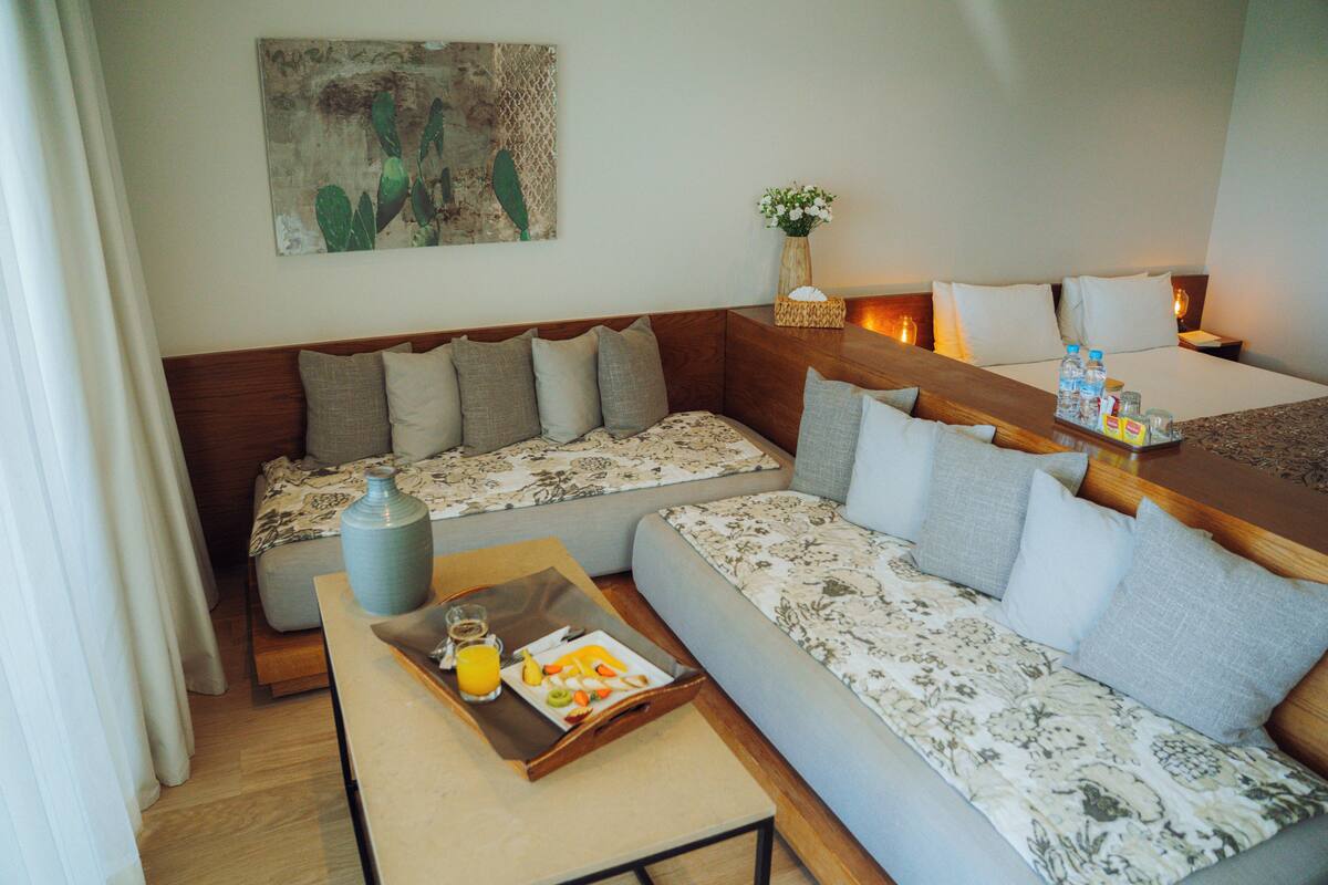 A hotel room with a sofa, a small table with breakfast, a vase with flowers, and a painting on the wall.