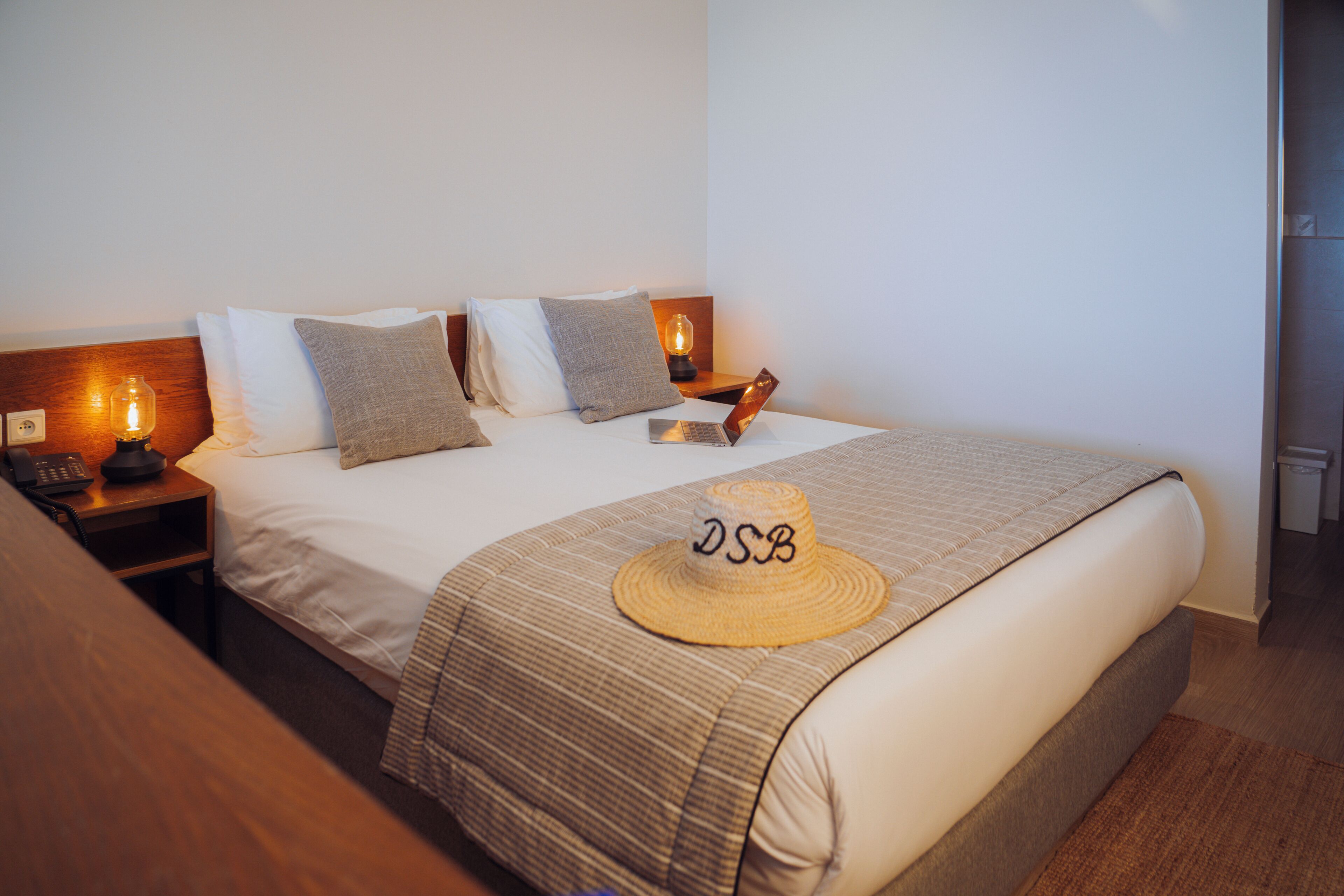 A hotel room with a neatly made bed, a bedside lamp, a hat on the bed, and a nightstand with a phone.