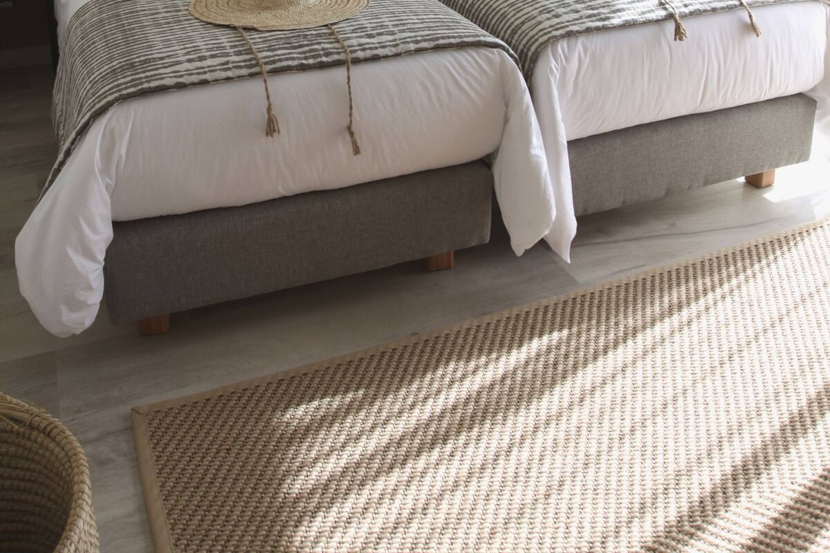 A hotel room with two beds, a nightstand, a lamp, and a woven rug.