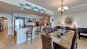 A spacious kitchen with a central island, bar stools, and a dining area with a large mirror and decorative lamps. - Dunes of Crystal Beach 104 | Free Seasonal Beach Service & More! (Destin)