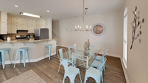 Una cocina moderna con armarios blancos, un refrigerador de acero inoxidable y un horno empotrado. Hay una zona de comedor con una mesa de vidrio y sillas azul claro. Un candelabro cuelga sobre la mesa y se observa un reloj de pared.