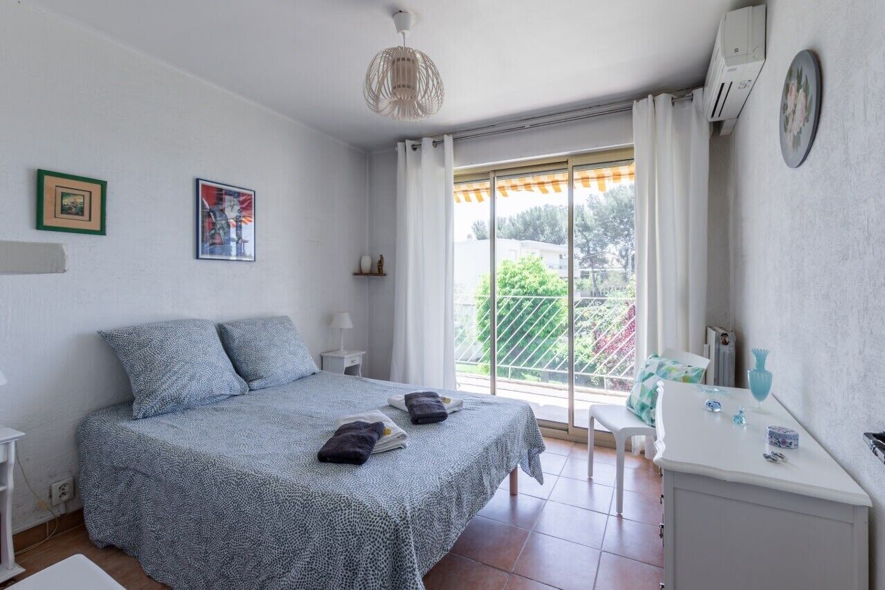 Un dormitorio con cama, escritorio, silla, ventana con cortinas y balcón.