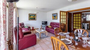 Una sala de estar con televisor, sofás rojos, una mesa de comedor para cuatro personas y una cocina visible a través de una puerta abierta.