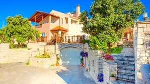 A two-story house with a white garage door, surrounded by stone walls and potted plants. - Villa Korini Crete (Mylopotamos)