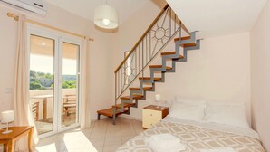A bedroom with a bed, a nightstand, a lamp, a wooden chair, a table, a staircase, and a sliding glass door leading to an outdoor area. - Villa Korini Crete (Mylopotamos)