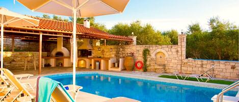 A swimming pool with lounge chairs and umbrellas, a stone wall, and a tiled roof structure.