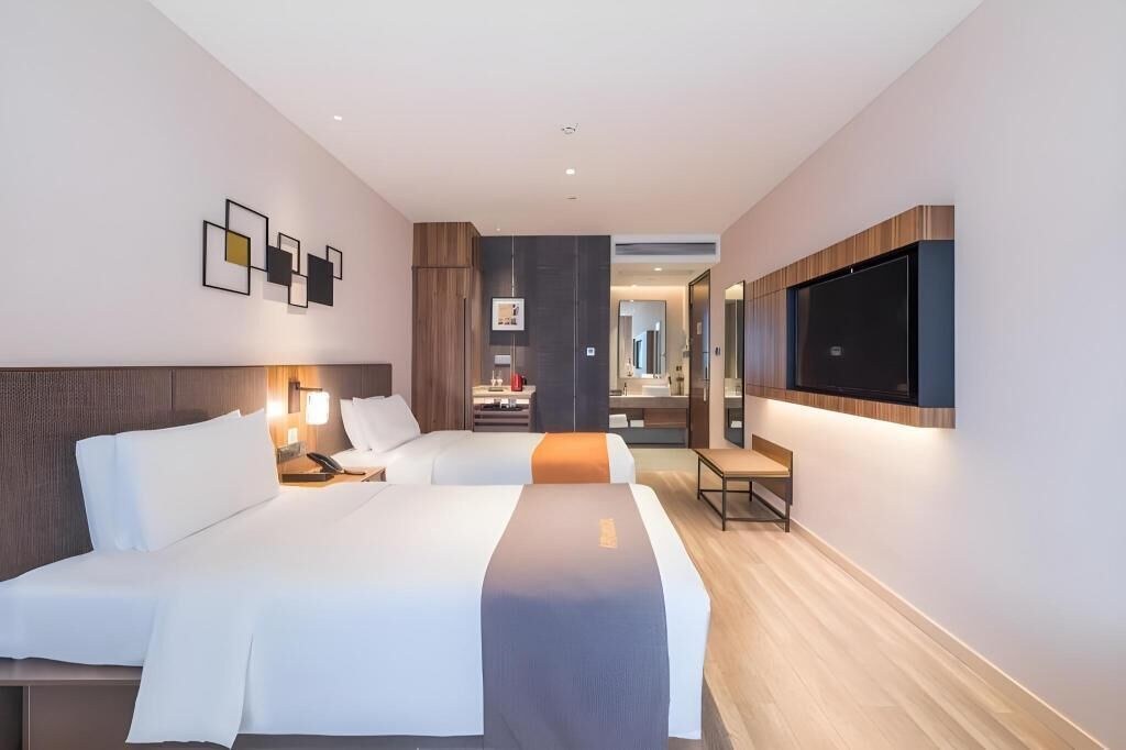 A modern hotel room with two beds, a TV, a desk, and a bathroom visible through an open door.