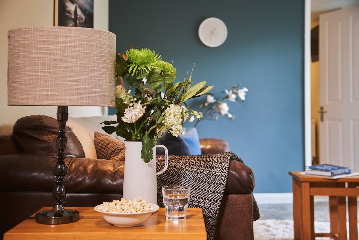 A living room with a brown leather sofa, a white vase with flowers, a lamp, a bowl of popcorn, and a glass of water on a wooden side table.