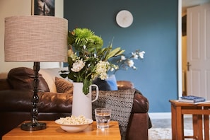 A living room with a brown leather sofa, a white vase with flowers, a lamp, a bowl of popcorn, and a glass of water on a wooden side table.