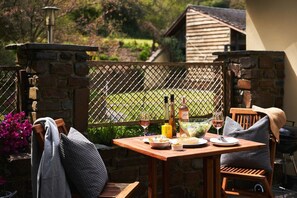 An outdoor dining area with a table set for two, a grill, and a view of a garden. - Swifts Return (Lower Ashton)