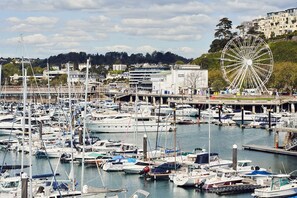 A marina with numerous boats docked, a large Ferris wheel in the background, and buildings on a hillside. - The Moorings (Torquay)