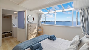 Een slaapkamer met een bed, een dressoir, een spiegel en uitzicht op zee door grote ramen.