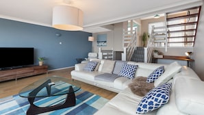 Een moderne woonkamer met een flatscreen televisie, een glazen salontafel, een witte bank met patroon kussens, en een trap die leidt naar een vide met boekenplanken.