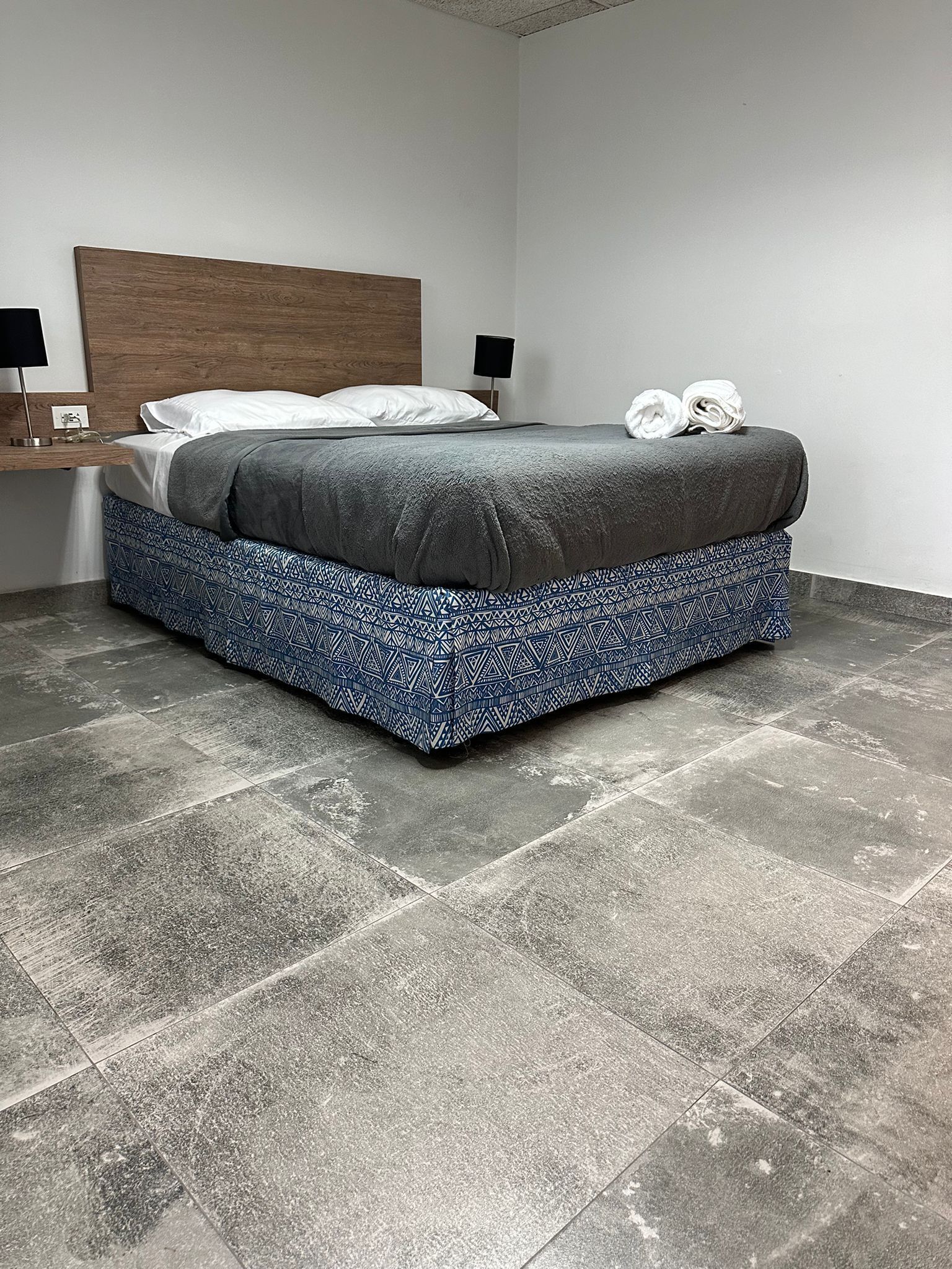 Quarto moderno com uma cama grande, cabeceira de madeira, mesa de cabeceira com abajur e saia de cama estampada.
