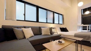 A modern hotel room with a sofa, a bed, a small table with a decorative item, and a large window with blinds. - b hotel Neko Yard | Stylish Loft Studio Central (Hiroshima City)