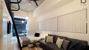 Une chambre d’hôtel moderne avec un lit, un canapé et une petite table. On y trouve un ventilateur de plafond, un radiateur et une fenêtre avec des stores.