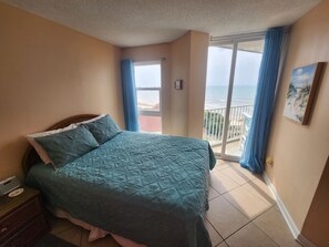 Un dormitorio con cama, mesita de noche, ventana con vistas a la playa y balcón con cortinas azules.