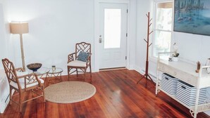 A room with hardwood flooring, a white door, a round woven rug, a wooden chair with a cushion, a small round table with a bowl and a candle, and a white console table with storage bins. - Modern Chic 1-BR Unit in the Cottages on James (Clayton)