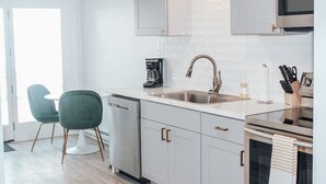 A modern kitchen with white cabinets, a stainless steel sink, and a coffee maker. There is a dining area with a round table and two chairs. - Modern Chic 1-BR Unit in the Cottages on James (Clayton)