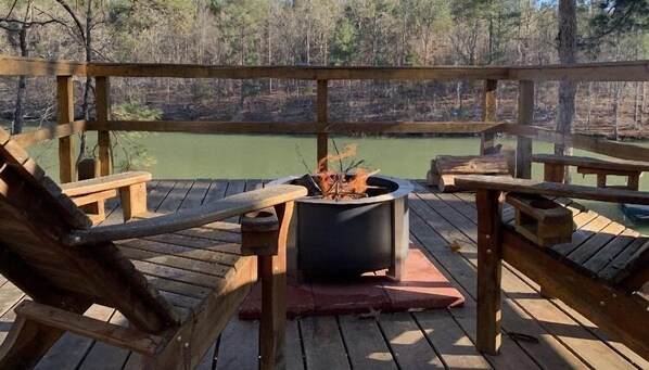 Enjoy a peaceful moment on the deck overlooking the serene water, surrounded by a forested backdrop and a cozy fire pit.