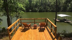 Une terrasse en bois avec deux chaises et une table, donnant sur un lac où se trouve un quai et un espace de repos abrité.