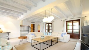 Ein geräumiges Wohnzimmer mit hoher Decke, einem Holztisch, einer Sofagarnitur, einem Fernseher und einer Treppe, die zu einer oberen Etage führt.