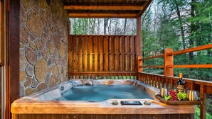 A hot tub with a stone wall and wooden deck, surrounded by a forest. - Spacious 5 BR/Triple Deck/View from top/close to all attractions in Gatlinburg. (Gatlinburg)