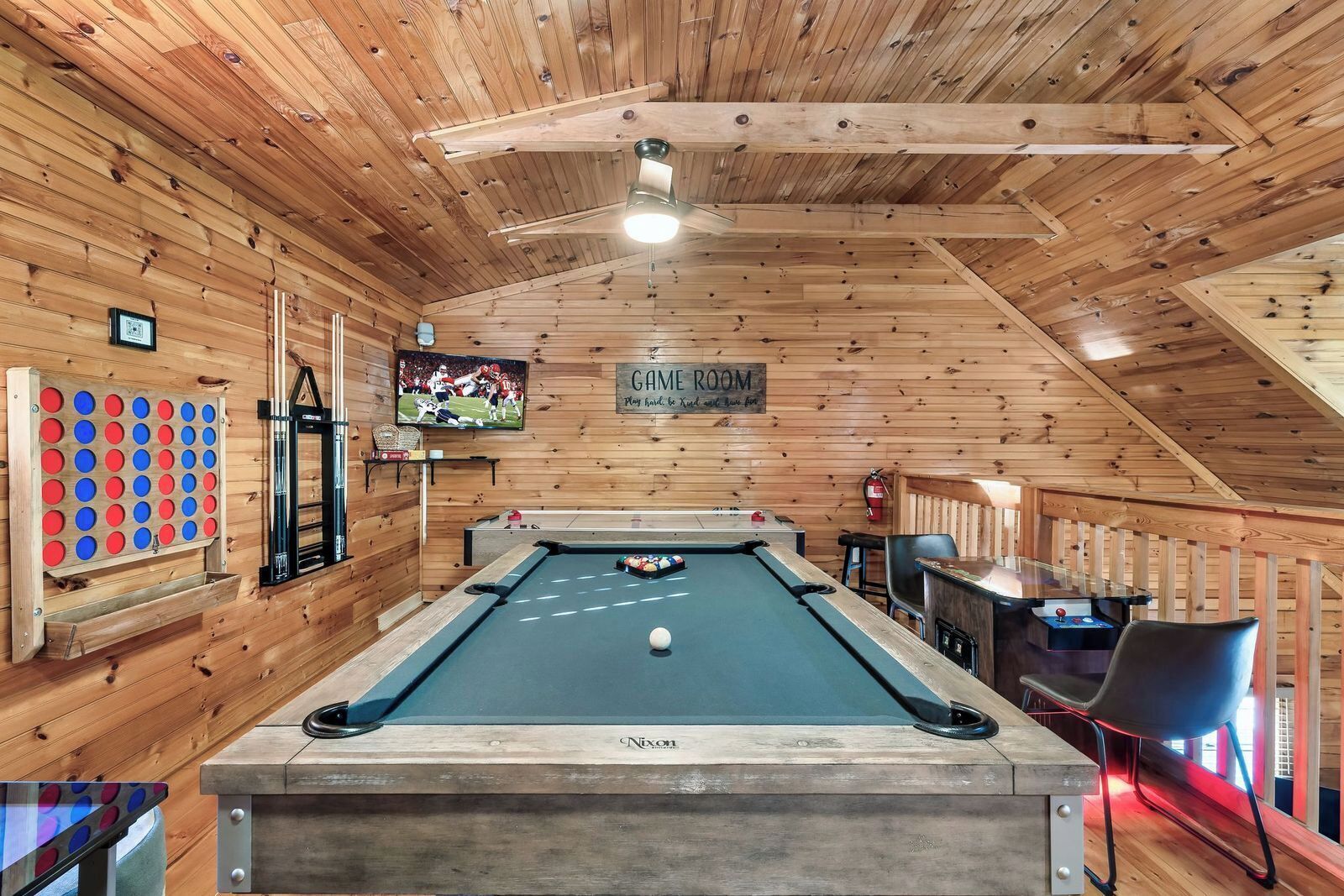 Een pooltafel in een houten kamer met een plafondventilator, een televisie en een bordje met de tekst 'GAME ROOM'.