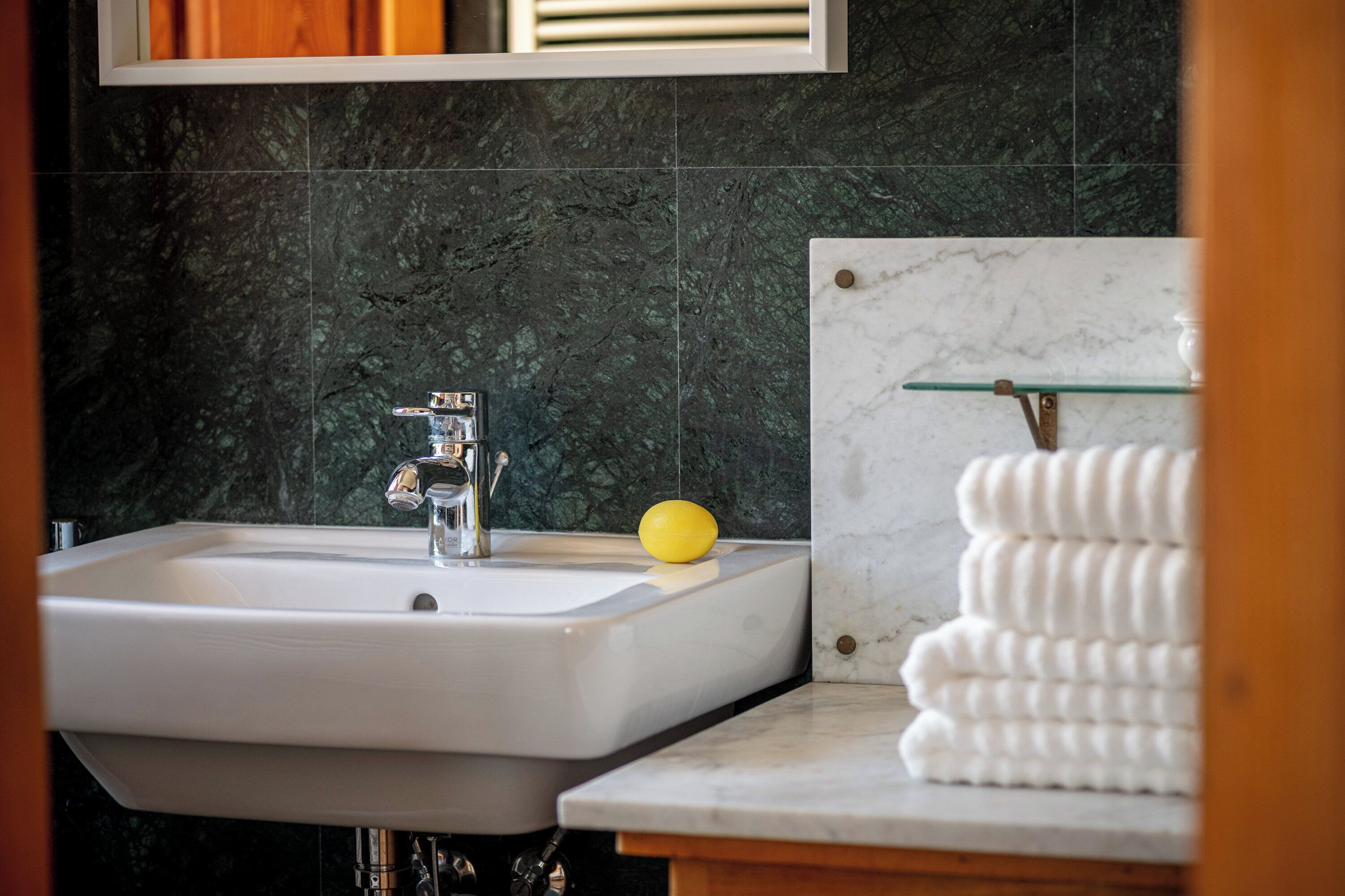A bathroom with a white sink, dark marble walls, and a glass shelf with towels.