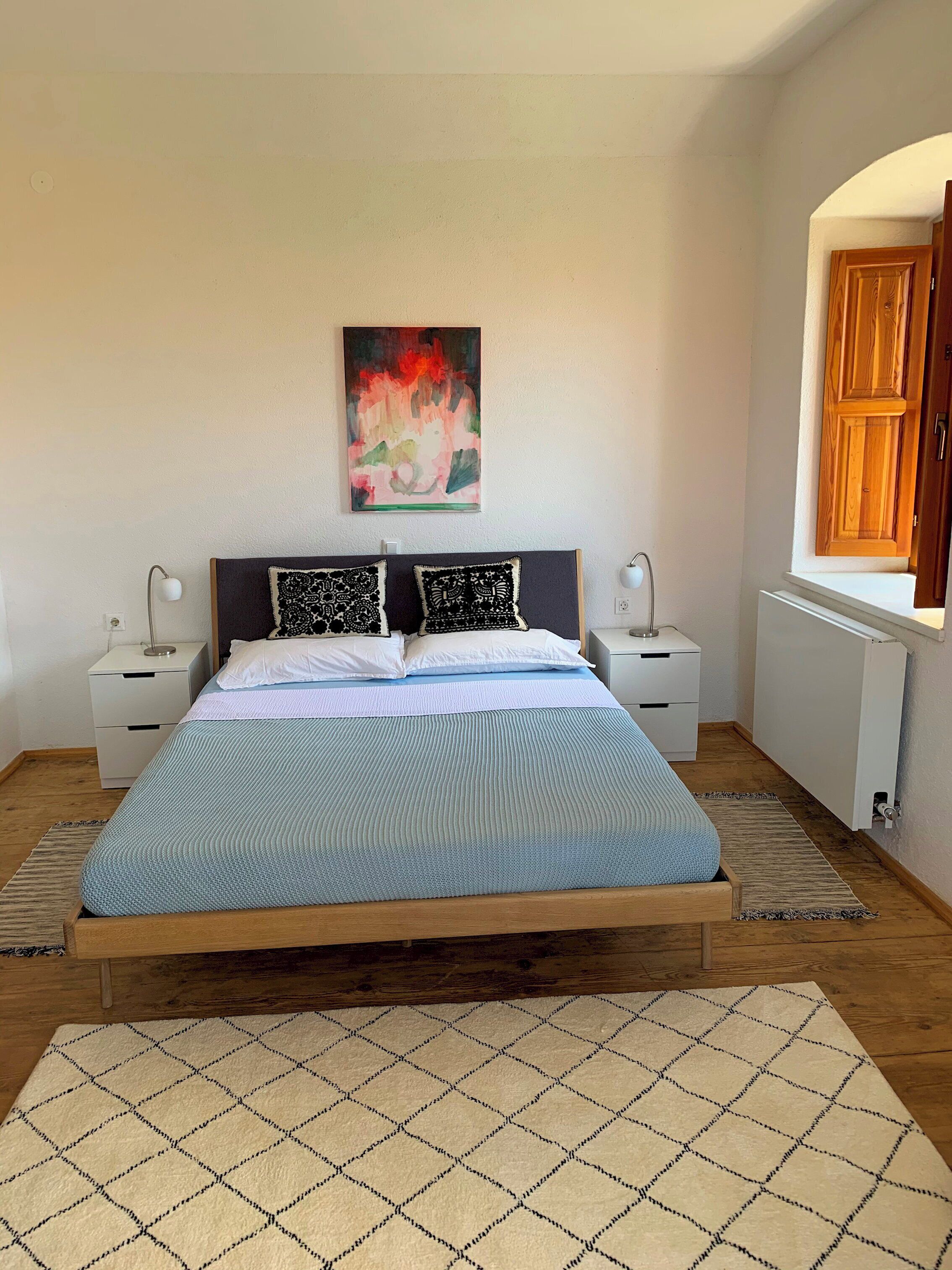 A bedroom with a bed, two bedside tables, a nightstand, a radiator, and a window with a wooden door.