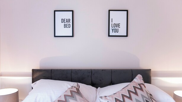 Un lit bien fait, avec deux oreillers et un couvre-lit à motifs. Deux panneaux encadrés sont accrochés au mur au-dessus du lit : l’un porte l’inscription « CHER LIT » et l’autre, « JE T’AIME ».