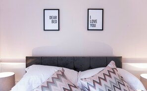 Una cama bien hecha con dos almohadas y un edredón estampado. Hay dos letreros enmarcados en la pared sobre la cama, uno dice "DEAR BED" y el otro "I LOVE YOU".