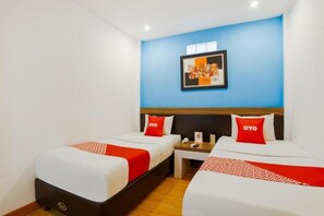 A hotel room with two beds, each with a red and white bedspread and a pillow with the word "OYO" on it. There is a small table between the beds with a phone and a sign.