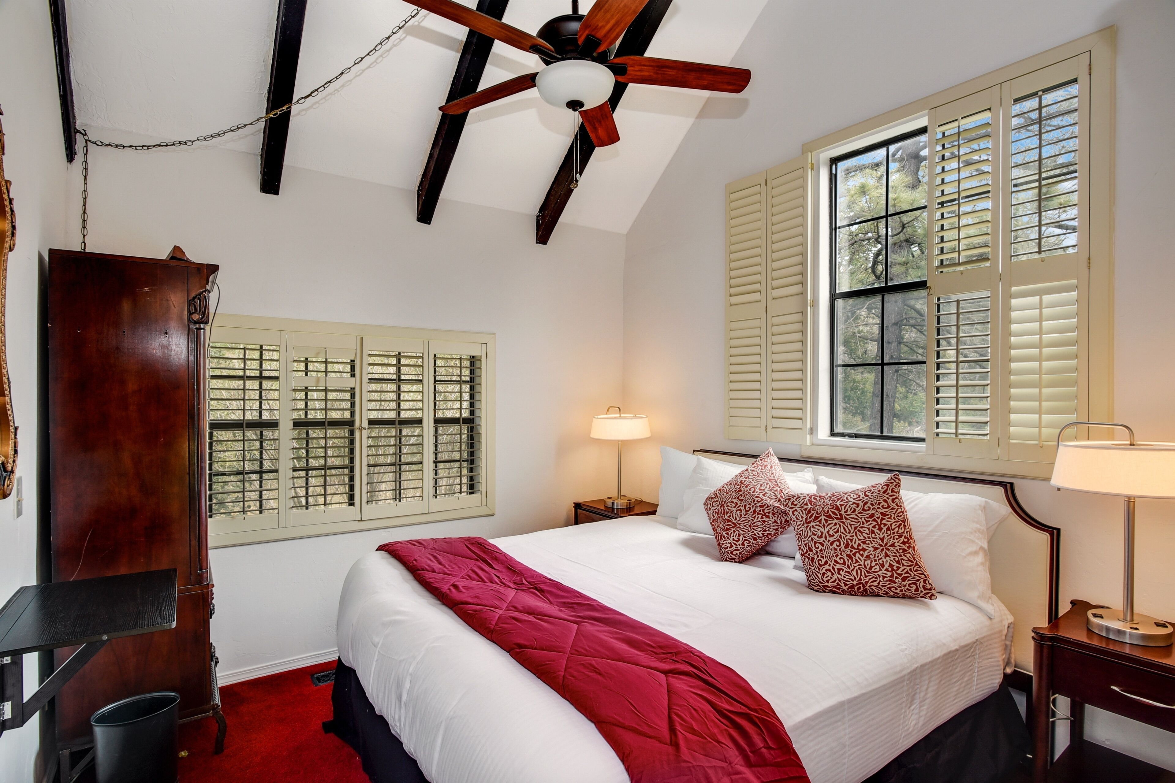 Une chambre d’hôtel comprenant un lit, un ventilateur de plafond, une fenêtre avec des volets, une table de chevet avec une lampe et une armoire en bois.