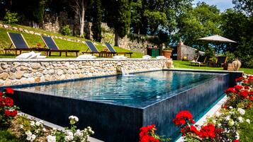 A rectangular swimming pool with a stone wall surrounding it, equipped with lounge chairs and a small table.