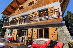 A two-story wooden chalet with a stone facade, a balcony with wooden railings, and outdoor seating. - CHALET FOR 16–18 PEOPLE WITH JACUZZI, SAUNA, MOVIE ROOM, AND VIEWS OF THE SLOPES AND MOUNTAINS (Montgenèvre)