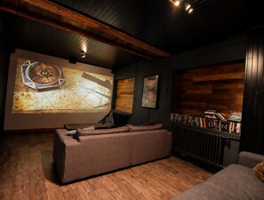 Una habitación acogedora con suelo de madera, una pantalla grande mostrando un mapa, una estantería y una zona de estar cómoda.