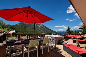Un área de comedor al aire libre con una sombrilla roja, cubierta de madera y vistas a la montaña.