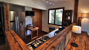 A modern kitchen with a stainless steel refrigerator, a wooden table, a gas stove, and a window providing a view of greenery outside.