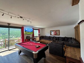 Una habitación con mesa de billar, sofá y televisor mostrando un paisaje de montaña.