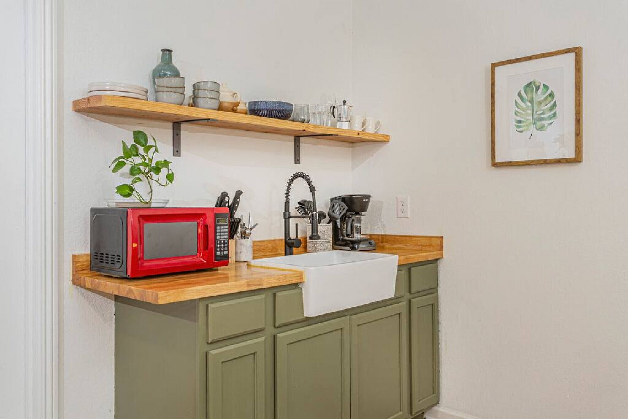 Une cuisine avec un micro-ondes rouge, un évier et une étagère en bois au-dessus, où sont rangés divers plats et une plante.