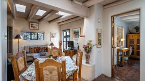 A cozy living room with a wooden dining table, chairs, and a sofa. There is a TV, a vase with flowers, and artwork on the walls.