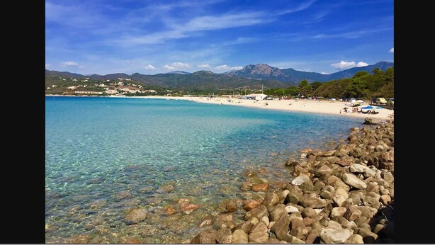 Une plage bordée d’une eau turquoise et limpide, d’une côte rocheuse et dominée par des montagnes en arrière-plan.