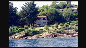 Un bâtiment situé sur la côte, doté d’une façade en pierre, de fenêtres en arc de cercle et d’un petit jardin avec des chaises.