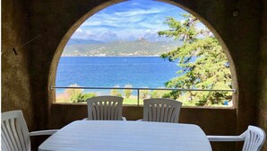Une table entourée de quatre chaises, dans une pièce semi-ouverte offrant une vue sur la mer et les collines.