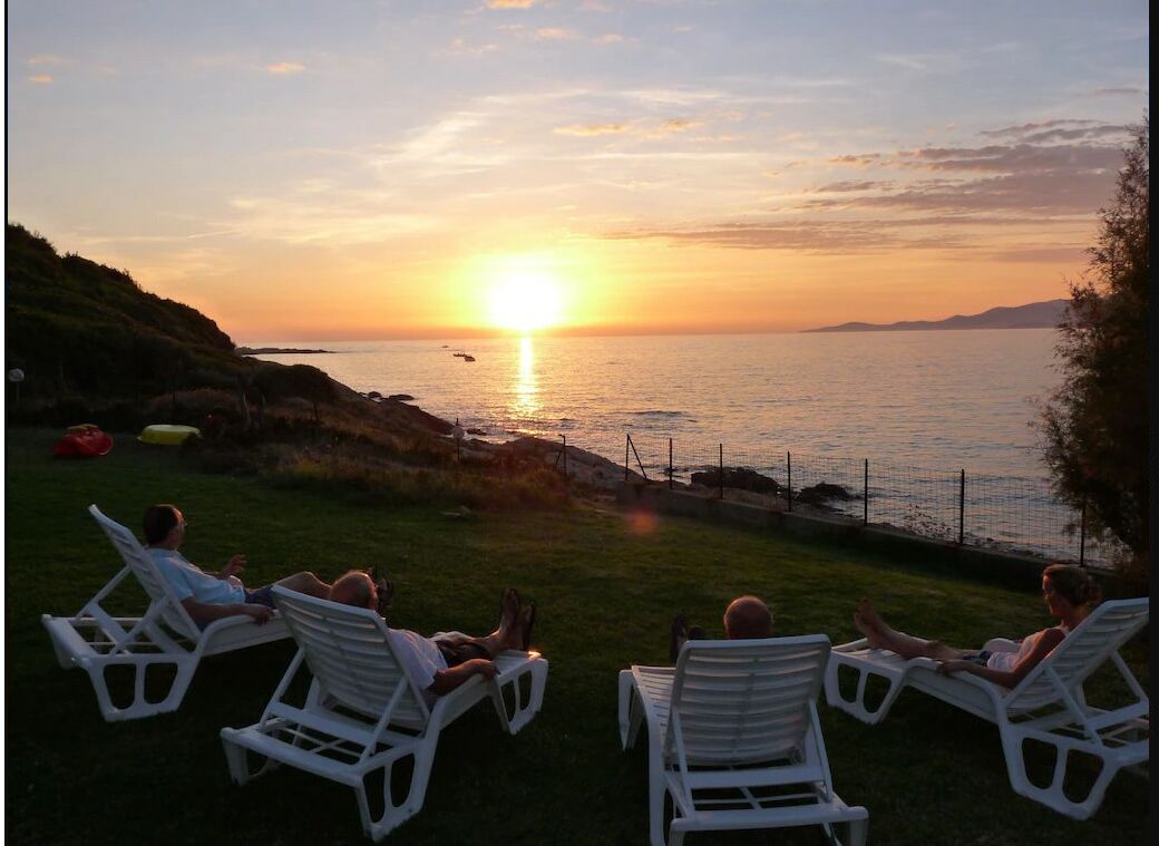 Une vue sur le coucher de soleil, prise depuis une zone herbeuse où se trouvent des chaises longues blanches, toutes orientées vers la mer.
