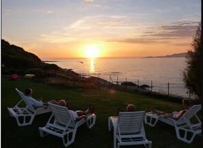 Une vue sur le coucher de soleil, prise depuis une zone herbeuse où se trouvent des chaises longues blanches, toutes orientées vers la mer.