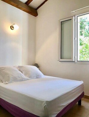 A bedroom with a bed, a nightstand, a wall-mounted light, and a window with a view of greenery. - Appartement vue mer et Montagne, à Létage (Calcatoggio)