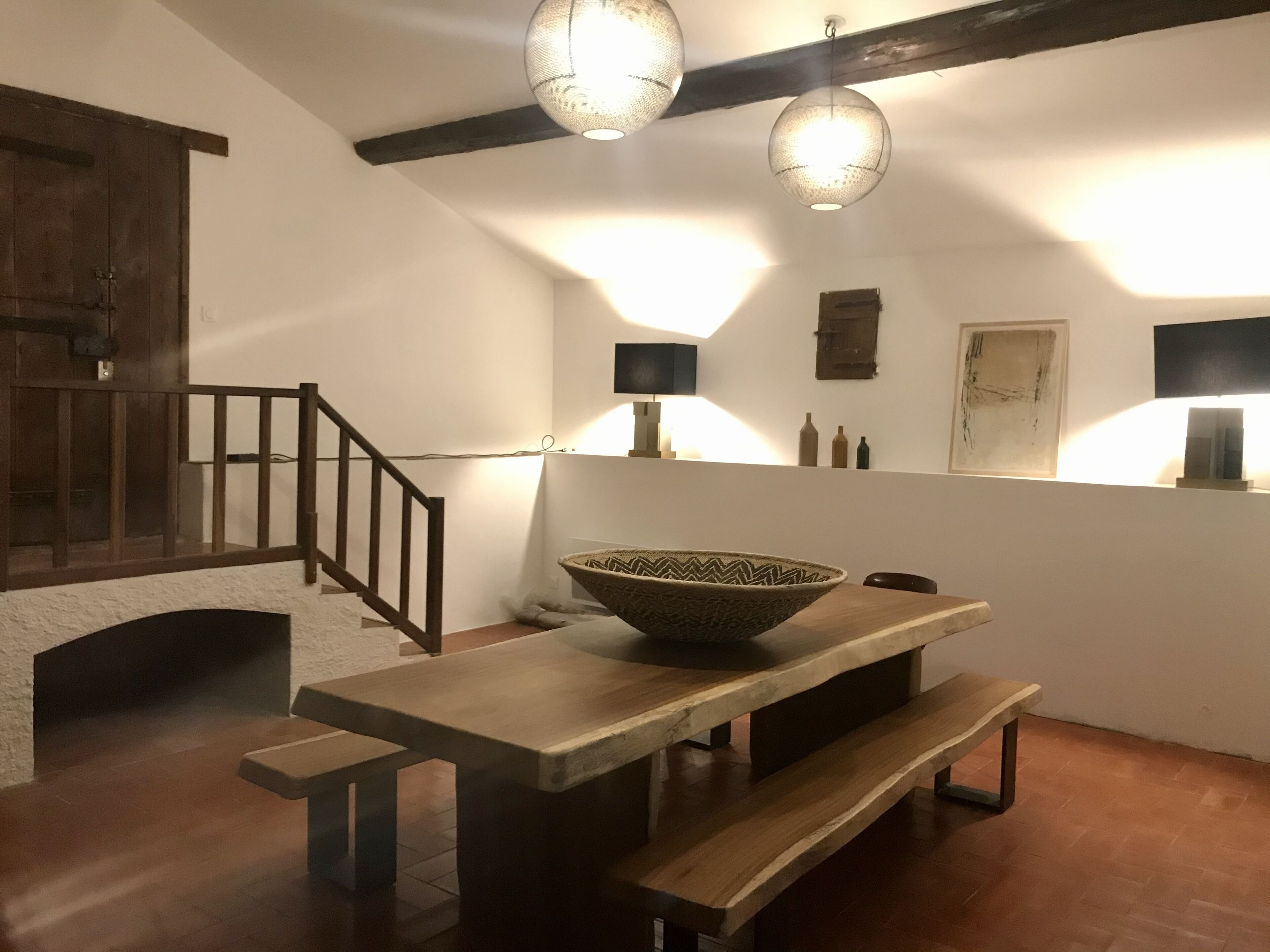 A dining area with a wooden table, benches, and a large woven bowl. There is a staircase with wooden railings and a wall-mounted shelf with bottles.