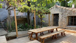 Une terrasse en bois avec une table de pique-nique et des bancs, entourée de murs en pierre et de verdure.